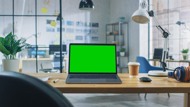 Laptop on the Desk in the Office Shows Green Mock-up Screen. In the Background Creative Modern Open Space Loft Office HUB with Professional Working