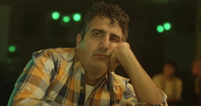 Drunk man leans on his elbow on a bar counter and looks sadly into the distance. He looks around, alute the others from the bar and drinks from his glass.