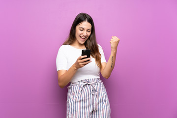 Young woman over isolated purple background with phone in victory position