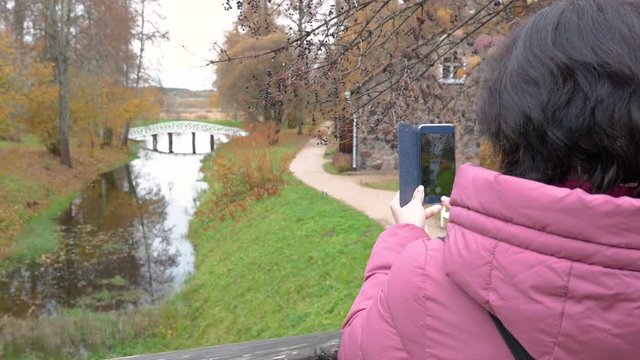 Cloudy autumn day in the background pond white bridge Caucasian middle aged woman brunette in glasses Burgundy down jacket takes a photo of nature on a smartphone