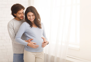 Loving man hugging his pregnant wife from behind standing near window