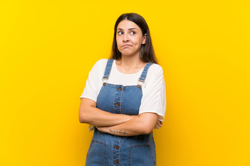 Young woman in dungarees over isolated yellow background making doubts gesture while lifting the shoulders