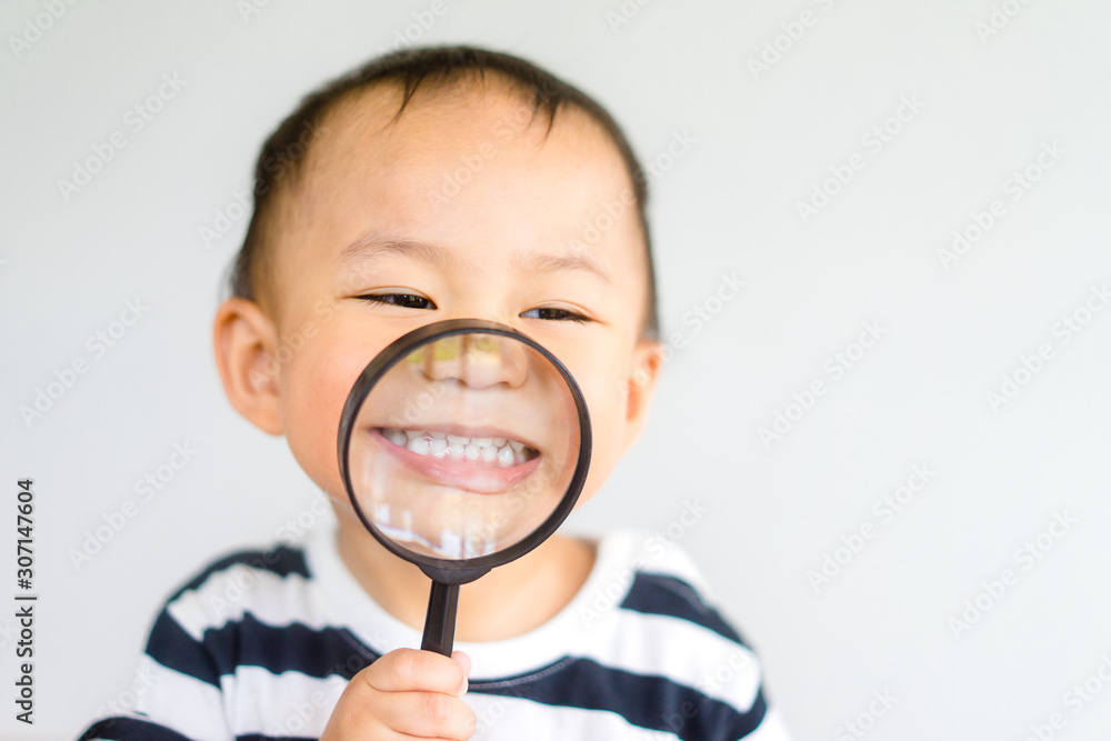 Wall mural Little toddler boy child holding magnifying glass and showing front teeth with big smile on green grass: Healthy happy funny smiling face young adorable lovely boy kid with new tooth dental.