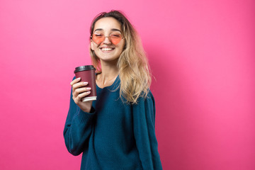 Glamor woman in glasses in a blue sweater with a drink of coffee on a pink background.