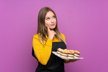 Teenager girl holding lots of different mini cakes over isolated purple background thinking an idea