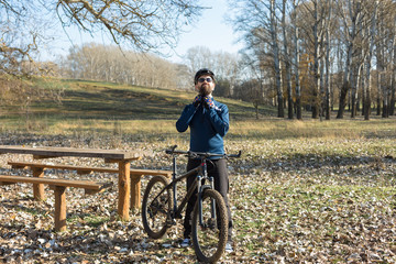 Cyclist in pants and fleece jacket on a modern carbon hardtail bike with an air suspension fork rides off-road. The guy is resting on a bench in the autumn park.