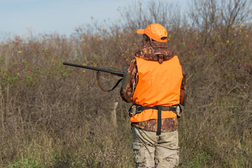 A man with a gun in his hands and an orange vest on a pheasant hunt in a wooded area in cloudy...
