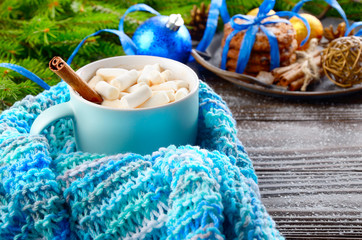 Christmas background of blue hot chocolate mug with marshmallows in knitted scarf, spruce branch and tray with gingerbread cookies on wooden table
