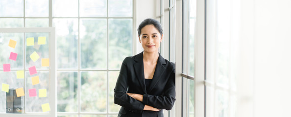 Portrait of smiling pretty young business woman crossed her arms and standing on workplace office and looking out to window.Smile and Thinking for future.