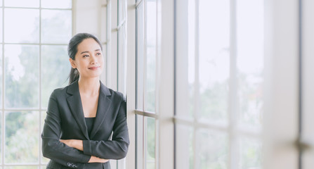 Portrait of smiling pretty young business woman crossed her arms and standing on workplace office and looking out to window.Smile and Thinking for future.
