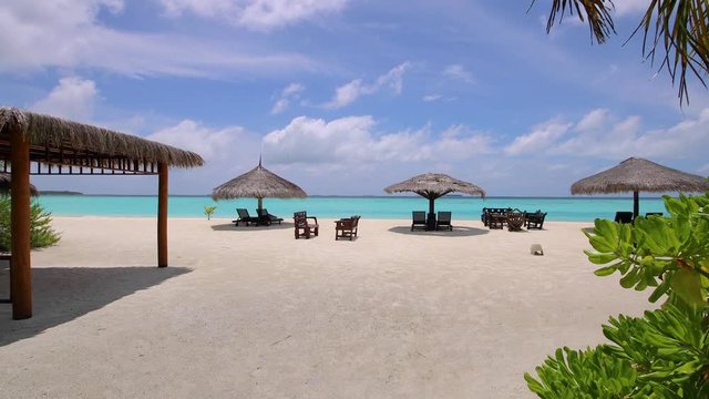 Beautiful shot of the entrance discovering a sandy beach in the Maldives. Palm trees, exotic trees, shrubs, white sand, shed. Close to the water beach chairs with umbrellas. Shot in forward motion