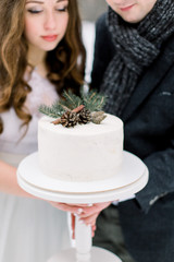 A wedding couple holds a beautiful white wedding cake, decorated with pine branch, cones and cinnamone, standing over the background of winter forest