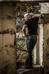 Bald brutal young man with tattoos stuffed on his arms and neck against the background of an abandoned destroyed building. Attractive tattooed stern guy. Concept of a bully and a dangerous guy.