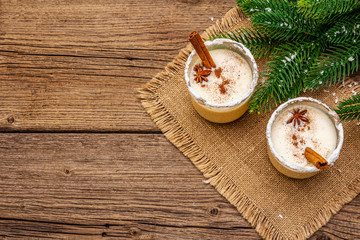 Eggnog cocktail in two glasses arranged with christmas decoration on old wooden table. Evergreen fer tree branch, artificial snow, sackcloth napkin