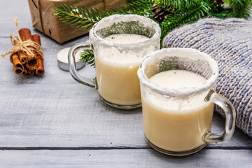 Eggnog cocktail in two glasses mug arranged with christmas decoration on light wooden table. Evergreen fir tree branch, gift, cozy jersey, artificial snow