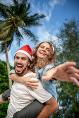 Fototapeta premium Young beautiful couple in Santa Claus hats walks along the shore of the tropical sea at the resort, vacation and travel for the New Year and Christmas time