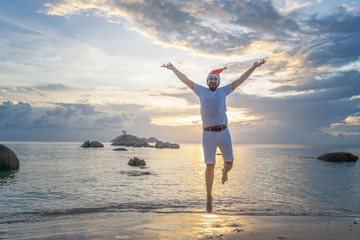 Young happy handsome latin man with beard in santa claus hat at tropical resort joyfully jumping, new year and christmas holidays and travel.