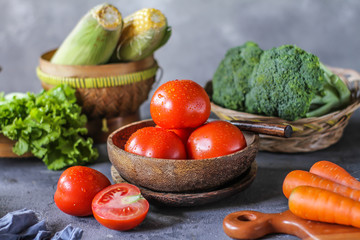 Photo of fresh tomatoes in a bowl on dark background around vegetables, carrot, salt, black pepper, corn, broccoli. Slice tomatoes. Harvesting tomatoes. Drops of water vegetables. Wooden table. Image