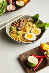 traditional asian ramen in bowl near chopsticks, vegetables on grey surface
