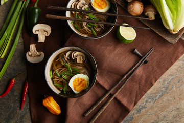 top view of traditional spicy ramen in bowls with chopsticks and vegetables on brown napkin on...