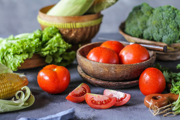 Photo of fresh tomatoes in a bowl on dark background around vegetables, carrot, salt, black pepper, corn, broccoli. Slice tomatoes. Harvesting tomatoes. Drops of water vegetables. Wooden table. Image