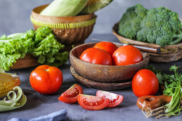 Photo of fresh tomatoes in a bowl on dark background around vegetables, carrot, salt, black pepper, corn, broccoli. Slice tomatoes. Harvesting tomatoes. Drops of water vegetables. Wooden table. Image