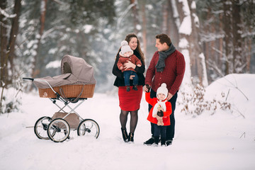 Fototapeta na wymiar A beautiful family with retro pram walks through the winter snowy forest. Mother, father, daughter and baby son enjoying day outdoors. Holidays, christmas, happiness together, childhood in love.