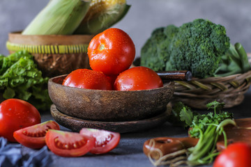 Photo of fresh tomatoes in a bowl on dark background around vegetables, carrot, salt, black pepper, corn, broccoli. Slice tomatoes. Harvesting tomatoes. Drops of water vegetables. Wooden table. Image