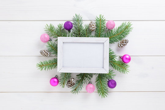 Photo frame between Christmas decoration, with pink balls and pine cones on a white wooden table. Top view, frame to copy space