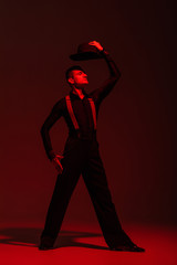 stylish dancer holding hat above head while performing tango on dark background with red lighting