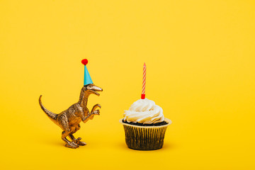 Toy dinosaur in party cap and cupcake with candle on yellow background