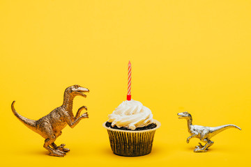 Toy dinosaurs beside cupcake with candle on yellow background