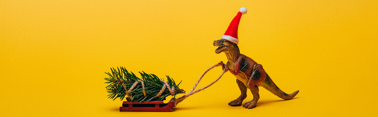Panoramic shot of toy dinosaur in santa hat with pine on sleigh on yellow background
