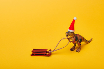 Toy dinosaur in santa hat with sleigh on yellow background