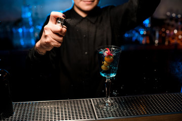 Female bartender spraying on the cocktail in the glass with berries and flower on the skewer with a bitter