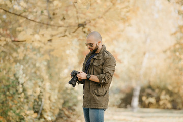 Bald photographer with a beard in aviator sunglasses with mirror lenses, olive cargo military jacket, blue jeans and shirt with digital wristwatch scrolls photos on the camera in the forest.