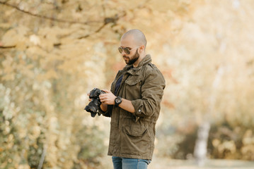 Bald photographer with a beard in aviator sunglasses with mirror lenses, olive cargo military jacket, blue jeans and shirt with digital wristwatch scrolls photos in the camera in the forest.