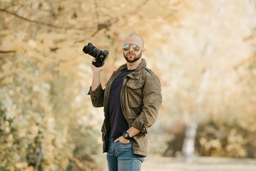 Bald photographer with a beard in aviator sunglasses with mirror lenses, olive cargo military jacket, blue jeans and shirt with digital wristwatch holds the camera in a bent hand in the forest.