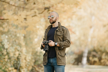 Bald photographer with a beard in aviator sunglasses with mirror lenses, olive military combat jacket, blue jeans and shirt with digital wristwatch holds the camera and looks straight in the forest.