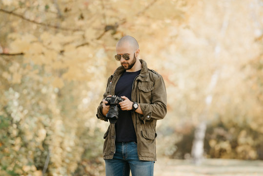 Bald photographer with a beard in aviator sunglasses with mirror lenses, olive cargo military jacket, blue jeans and shirt with digital wristwatch checks photos in the camera in the forest.