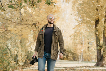 Fototapeta na wymiar Bald photographer with a beard in aviator sunglasses with mirror lenses, olive military jacket, jeans and shirt with digital wristwatch holds the DSLR camera and goes straight on the battlefield 