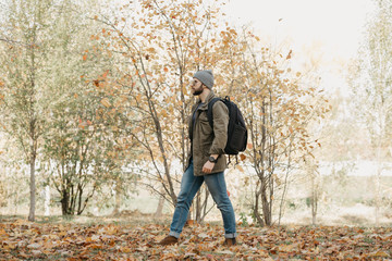 A brutal traveler with a beard in aviator sunglasses with mirror lenses, olive military combat jacket, jeans, hat with backpack and wristwatch goes through the forest in the noon. A photojournalist.