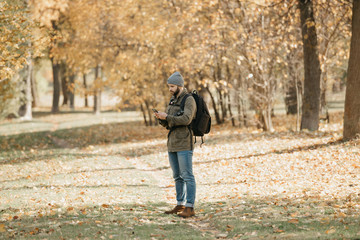 A traveler with a beard in olive military cargo combat jacket, jeans, hat with backpack and wristwatch holds the DSLR camera and checks for something in his smartphone.