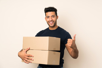 Young handsome man over isolated background holding a box to move it to another site with thumb up