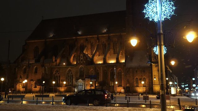 Beautiful Snowfall at night. White Winter in Wroclaw, Poland. St. Mary Magdalene Church Christmas lights moving cars and taxi