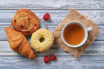 Sweet yellow donut with white sprinkles, puff bun with cream and raisins in the shape of a snail and fresh croissants on blue glazed saucer. Dessert with cap of tea, berries of fresh juicy raspberries