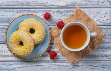 Sweet yellow donuts with white sprinkles on blue glazed saucer. Dessert with tasty, delicious doughnuts, cup of green chamomile tea, fresh juicy red raspberries on natural wooden background.Sweet yell
