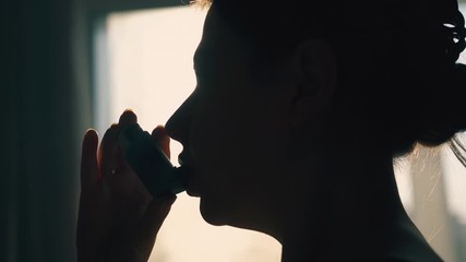 CU: black silhouette of young woman using inhaler to cure asthma at backlight near window in room extreme close view