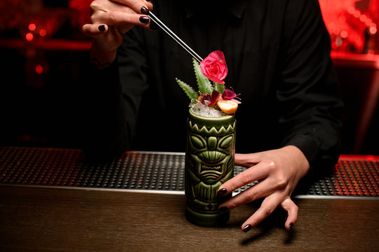 Bartender girl serving alcoholic cocktail in the Tiki mug adding a rose bud with tweezers