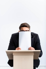 scared businessman in suit standing at podium tribune and obscuring face with paper during...